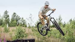 Ein Mountainbiker mit Helm springt mit einem Root Miller 4 Mountainbike über ein Hindernis in einer natürlichen, bewaldeten Umgebung.