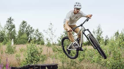 Ein Mountainbiker mit Helm springt mit einem Root Miller 4 Mountainbike über ein Hindernis in einer natürlichen, bewaldeten Umgebung.