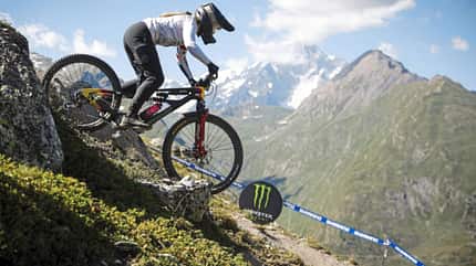 Enduro-Rennen, Mountainbike, La Thuile, MTB World Series, Alpen, Downhill, Fahrradrennen, Shimano, Monster Energy, Berglandschaft Mountainbikerin fährt bei einem Enduro-Rennen in alpiner Umgebung in La Thuile einen steilen Trail hinab. Im Hintergrund sind Berge und ein Monster-Energy-Logo sichtbar.