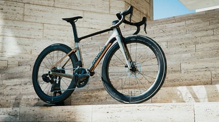 Aero-Rennrad, Hochprofil-Laufradsatz, Scheibenbremse, Rennlenker, Kettenschaltung, Straßenfahrrad, Außenaufnahme, Architekturhintergrund, Continental-Reifen Das Foto zeigt ein modernes Aero-Rennrad mit tiefen Aero-Laufrädern und Dropbar-Lenker, seitlich auf hellen Steinstufen positioniert. Sichtbar sind Scheibenbremsen, ein aufgeräumtes Cockpit mit interner Kabelführung und eine klassische Kettenschaltung; die Reifen tragen Continental-Schriftzüge. Die Aufnahme entstand im Außenbereich vor einer zeitgenössischen Architekturkulisse.
