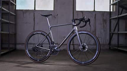 Rennrad, Scheibenbremse, Aero-Laufrad, Dropbar, Zweifach-Kettenblatt, Polymer-Laufrad, Straßenfahrrad, Studioaufnahme, Fahrradtechnik Das Foto zeigt ein modernes Rennrad mit Dropbar-Lenker, Scheibenbremsen und tiefen Aero-Laufrädern mit dem Schriftzug „POLYMER“. Der Rahmen ist in vollständiger Seitenansicht dargestellt; rechts ist die Antriebsseite mit Zweifach-Kurbel und Kettenschaltung sichtbar. Die Aufnahme entstand in einem schlichten Innenraum mit Betonboden, großen Fenstern und Regalreihen im Hintergrund.