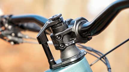 Fahrrad-Vorbau, Lenkerhöhenverstellung, Schnellspanner, Ahead-System, Vorbauklemme, Fahrradlenker, Steuersatz, Cockpit Fahrrad, Mountainbike-Komponente, Fahrradtechnik Detailaufnahme eines Fahrrad-Vorbaus mit Lenker an einer höhenverstellbaren Steuersäule mit Schnellspanner (Ahead-System). Sichtbar sind Vorbauklemme, Topcap und Kabelzüge; die schwarz eloxierten Komponenten bilden das Cockpit des Fahrrads. Die Szene ist im Freien aufgenommen, Hintergrund unscharf.