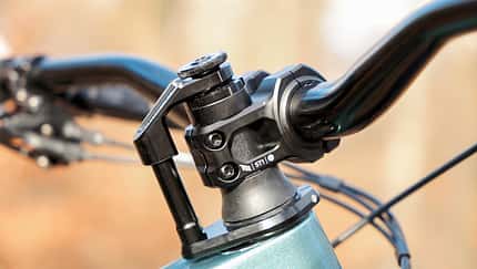 Fahrrad-Vorbau, Lenkerhöhenverstellung, Schnellspanner, Ahead-System, Vorbauklemme, Fahrradlenker, Steuersatz, Cockpit Fahrrad, Mountainbike-Komponente, Fahrradtechnik Detailaufnahme eines Fahrrad-Vorbaus mit Lenker an einer höhenverstellbaren Steuersäule mit Schnellspanner (Ahead-System). Sichtbar sind Vorbauklemme, Topcap und Kabelzüge; die schwarz eloxierten Komponenten bilden das Cockpit des Fahrrads. Die Szene ist im Freien aufgenommen, Hintergrund unscharf.