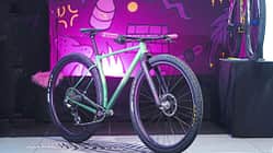 Ein modernes Mountainbike mit farbigem Rahmen und breiten Stollenreifen steht auf einer Ausstellungsfläche vor einer bunt beleuchteten Wand mit Comic-Grafik. Das Fahrrad ist komplett aufgebaut und wird prominent präsentiert.