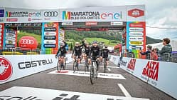 Gruppe von Radfahrern überquert die Ziellinie beim Maratona dles Dolomites 2024 unter dem offiziellen Zielbogen mit Sponsorenlogos.