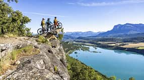 Drei Mountainbiker stehen mit ihren Fahrrädern auf einem Felsvorsprung oberhalb eines Sees mit Panoramablick auf Berge und Landschaft.