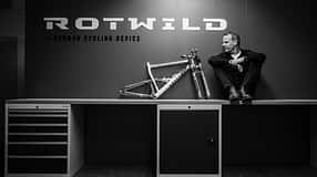 Schwarzweiß-Foto einer Person, die auf einer Werkbank neben einem Fahrradrahmen sitzt. Im Hintergrund das Logo 'Rotwild' und der Schriftzug 'German Cycling Device'.