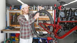 Person wartet ein vollgefedertes Mountainbike in einer Fahrradwerkstatt. Das Fahrrad ist in einem Montageständer eingespannt, im Hintergrund sind Werkzeuge und Ersatzteile sichtbar.