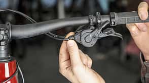 Detailaufnahme eines Fahrradlenkers mit Schalthebel und Kabel, an dem eine Hand arbeitet. Der Dateiname liefert keine spezifischen Modell- oder Markendaten, daher allgemeine Kategorisierung als Fahrradkomponente.