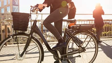 Low section of mature woman riding electric bicycle by commuters on bridge in city against sky