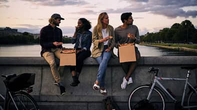 Vier Freunde essen Pizza auf einer Brücke bei Sonnenuntergang, mit Fahrrädern im Vordergrund.