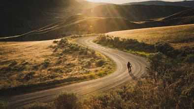 Gravelbiker fährt bei Sonnenuntergang eine staubige Serpentinenstraße durch hügelige Landschaft
Gravelbiker fährt bei Sonnenuntergang eine staubige Serpentinenstraße durch hügelige Landschaft