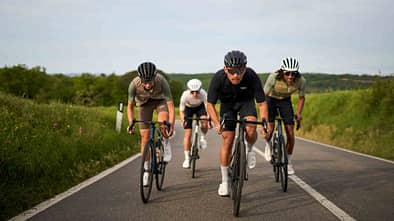 Drei Rennradfahrer fahren in Teamformation auf einer offenen Landstraße, flankiert von grüner Natur.