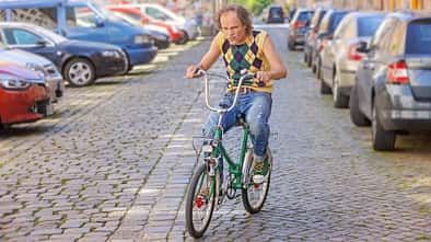 Olaf Schubert fährt ein Retro-Fahrrad auf einem Parkplatz