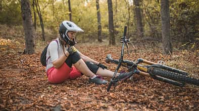 Eine Mountainbikerin sitzt in einem Wald mit herabgefallenem Herbstlaub, nachdem sie vom Fahrrad gefallen ist und sich verletzt hat.