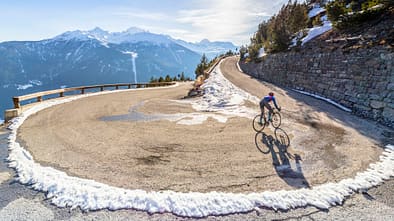 Road cyclist climbing hairpin bends up mountain pass in winter with snow.