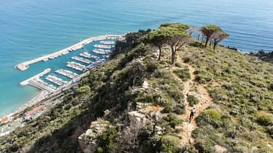 Luftaufnahme eines Küstenhügels mit Mountainbiker auf einem Trail, darunter ein Yachthafen am Meer.
