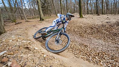 Mountainbiker in voller Schutzausrüstung fährt auf einem Trail im Wald durch eine Steilkurve.