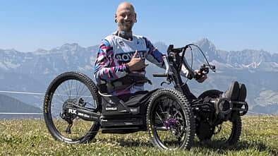 Eine Person sitzt in einem geländegängigen Handbike (Bowhead RX) auf einer Wiese vor Bergpanorama.