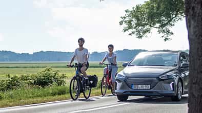 Zwei Radfahrer fahren auf einer Landstraße und werden von einem Auto überholt 