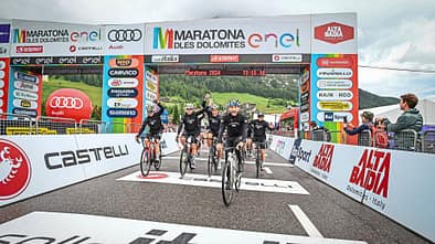 Gruppe von Radfahrern überquert die Ziellinie beim Maratona dles Dolomites 2024 unter dem offiziellen Zielbogen mit Sponsorenlogos.