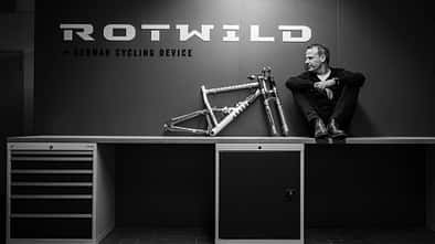 Schwarzweiß-Foto einer Person, die auf einer Werkbank neben einem Fahrradrahmen sitzt. Im Hintergrund das Logo 'Rotwild' und der Schriftzug 'German Cycling Device'.