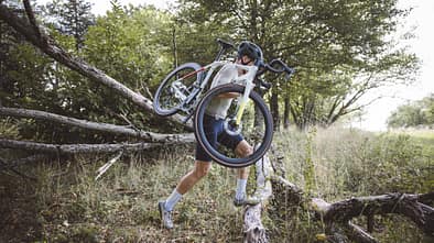 Das Foto zum Thema „Cyclo vs Gravel“ zeigt eine Person, die ein Dropbar-Geländerad (Cyclocross/Gravel-Bike) über umgestürzte Baumstämme in einem Wald trägt. Sichtbar sind Rennlenker, Scheibenbremsen und breite Stollenreifen – eine typische Tragepassage aus dem Cyclocross, wie sie auch beim Gravel-Einsatz vorkommt.