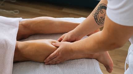 Close-up side view hands of unrecognizable male masseur with strong tattooed hands massaging lower leg of muscular athlete man after sports workout. Close-up side view hands of unrecognizable male masseur with strong tattooed hands massaging lower leg of muscular athlete man after sports workout.