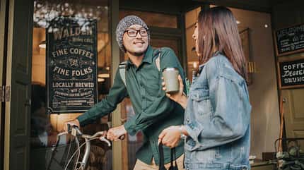Junger Mann mit Fahrrad spricht mit einer Frau vor einem Café, beide halten Kaffee in der Hand. Junger Mann mit Fahrrad spricht mit einer Frau vor einem Café, beide halten Kaffee in der Hand.
