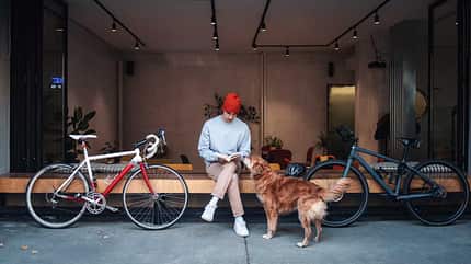 Junger Mann mit roter Mütze liest ein Buch vor einem Café, während ein Hund ihn anschaut. Zwei Fahrräder lehnen an der Bank. Junger Mann mit roter Mütze liest ein Buch vor einem Café, während ein Hund ihn anschaut. Zwei Fahrräder lehnen an der Bank.