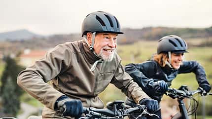 E-Bike, Senioren, Fahrradfahren, Outdoor, Gesundheit, Bewegung, Lebensfreude, Aktivität im Alter, Paar, Natur, Helm, Sport, Radfahren, Fitness, Freizeit, Abenteuer Älteres Paar fährt lächelnd mit E-Bikes durch hügelige Landschaft, beide mit Helmen und Outdoor-Bekleidung.