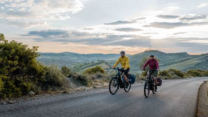 Bosch Performance Line PX, E-Bike Tour, Fahrradreise, Trekkingbike, Reiseradler, Sonnenuntergang, Packtaschen, Hügellandschaft, Fahrradtour, Bosch Motor, Fahrradabenteuer, Natur erleben, Bikepacking. Zwei Fahrradfahrer mit Packtaschen fahren auf einer asphaltierten Straße durch eine hügelige Landschaft bei Sonnenuntergang – Symbolbild für E-Bike-Touren mit Bosch Performance Line PX.