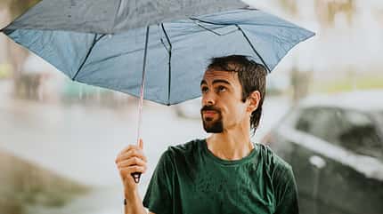 Ein genervter junger Mann wird mit einem kaputten Regenschirm vom Regen überrascht. Ein genervter junger Mann wird mit einem kaputten Regenschirm vom Regen überrascht.