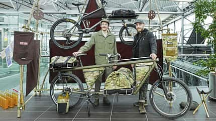 Zwei Männer präsentieren ein Lastenfahrrad mit Tarnmuster-Taschen auf einer Indoor-Ausstellung. Im Hintergrund ist ein weiteres Fahrrad auf einem Holzgestell zu sehen. Die Umgebung ist modern und lichtdurchflutet.