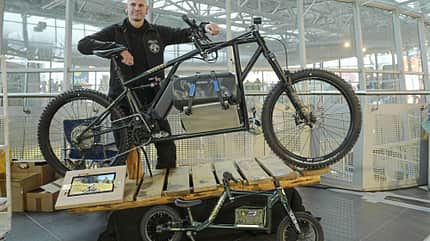 Ein Mann präsentiert ein großes, individuell aufgebautes E-Bike mit Lastenrahmen auf einer Messeplattform. Vor dem E-Bike steht ein passendes Kinderlaufrad im ähnlichen Design. Im Hintergrund ist eine moderne Messehalle mit Glasdach zu sehen.