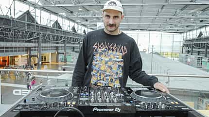 DJ an einem Pioneer DJ-Controller in einer modernen Messe- oder Bahnhofshalle, frontal aufgenommen.