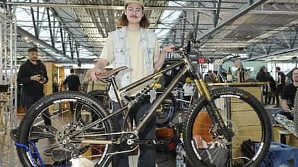 Eine Person präsentiert ein Mountainbike auf einer Fahrradmesse oder Ausstellung. Das Fahrrad steht auf einem Montageständer, im Hintergrund sind weitere Besucher und Aussteller zu sehen.