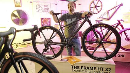 Ein Mann präsentiert ein Mountainbike-Prototyp 'THE FRAME HT 32' von Bike Ahead Composites auf einer Messe. Das Fahrrad steht im Vordergrund, im Hintergrund sind weitere Fahrradteile und Rahmen ausgestellt.