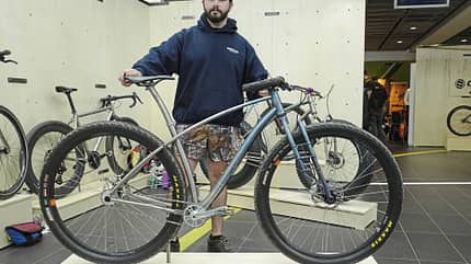 Ein Mann steht in einer Indoor-Ausstellungshalle hinter einem modernen Mountainbike mit dicken Reifen. Im Hintergrund sind weitere Fahrräder auf Podesten ausgestellt.