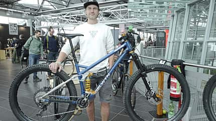 Ein Mann steht in einer Messehalle hinter einem modernen Mountainbike mit Vollfederung und breiten Reifen. Das Fahrrad trägt den Markennamen 'Selwyn' und ist mit einer RockShox Lyrik Federgabel sowie Schwalbe Reifen ausgestattet. Im Hintergrund sind weitere Personen und Fahrräder in einer modernen Ausstellungsumgebung zu sehen.