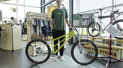 Person präsentiert ein gelbes Mountainbike vor einem Messestand mit weiteren Fahrrädern und Bekleidung.