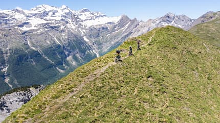 Mountainbike, Alpen, Berggrat, Singletrail, Outdoor, Gebirge, Fahrrad, Panorama, Hochgebirge, Abenteuer Drei Mountainbiker fahren auf einem schmalen Berggrat mit Blick auf schneebedeckte Alpen im Hintergrund.