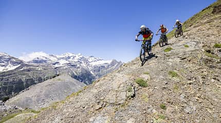 Mountainbike, Gebirgstour, Alpen, Fahrrad, Downhill, Outdoor, Sport, Fahrradhelm, Singletrail, Berglandschaft Drei Mountainbiker fahren auf einem steinigen Gebirgspfad mit schneebedeckten Bergen im Hintergrund.