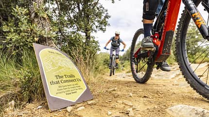 Mountainbike, E-Mountainbike, Trail, Schotterweg, Outdoor, Fahrrad, Zonazero, Bikepark, Naturlandschaft, Sport Zwei Mountainbiker fahren auf einem Schotter-Trail durch eine bewaldete Landschaft. Im Vordergrund ist ein Schild mit der Aufschrift 'The Trails that Connect Us' und Informationen zu den Trails zu sehen. Die Aufnahme zeigt ein modernes E-Mountainbike und einen weiteren Radfahrer im Hintergrund.