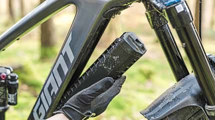 Detailaufnahme eines Giant E-Mountainbikes mit entnehmbarer Batterie im Unterrohr. Eine Person mit Handschuhen entnimmt den Akku aus dem Rahmen. Das Bike steht in einem Waldgebiet, der Fokus liegt auf dem Batteriewechsel.