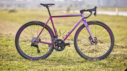 Das Bild zeigt ein lila-farbenes Rennrad mit ENVE-Laufrädern und Shimano Dura-Ace-Antrieb, aufgenommen im Freien auf einer Wiese mit unscharfem Stadt- und Brückenhintergrund. Der Dateiname weist auf ein Produktfoto des Modells IRIS x St Joris hin.