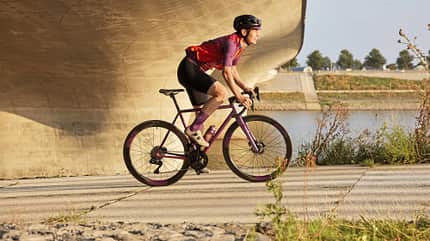 Fahrradfahrerin auf violettem Rennrad unter moderner Brücke an einem Flussufer, sommerliche Szene.