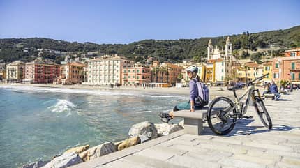 Person mit Fahrrad sitzt auf einer Bank an einer mediterranen Strandpromenade, bunte Häuser und Meer im Hintergrund.