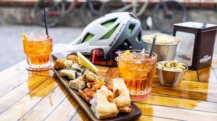 Auf einem Holztisch stehen zwei Gläser mit Aperitif, ein Tablett mit italienischen Snacks, ein Fahrradhelm der Marke Endura, Sonnenbrille, Schalen mit Chips und Gebäck. Im Hintergrund sind unscharf Fahrräder zu erkennen.