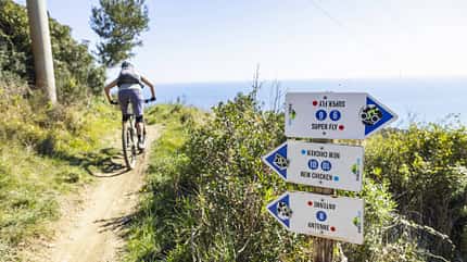 Mountainbiker auf einem Trail an der Küste, Wegweiser mit verschiedenen Trailnamen im Vordergrund.